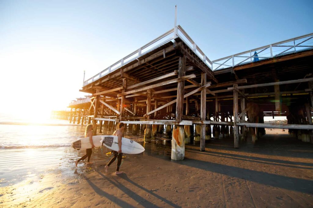 Two surfers with boards walking along the wet beach at sunset beneath a wooden pier.