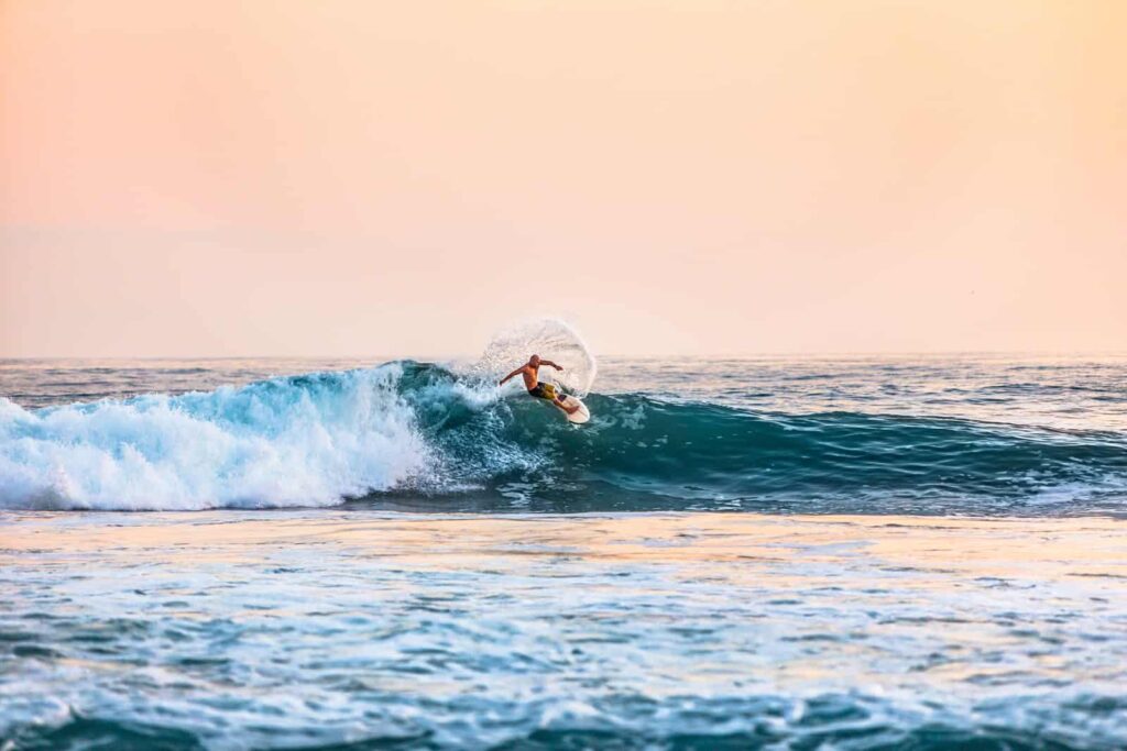 Surfer riding a curling blue wave at sunset with pink-orange sky in the background.