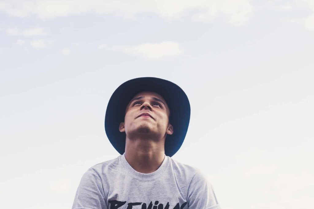 Young man wearing a wide-brim hat looks up at the sky outdoors on a cloudy day.