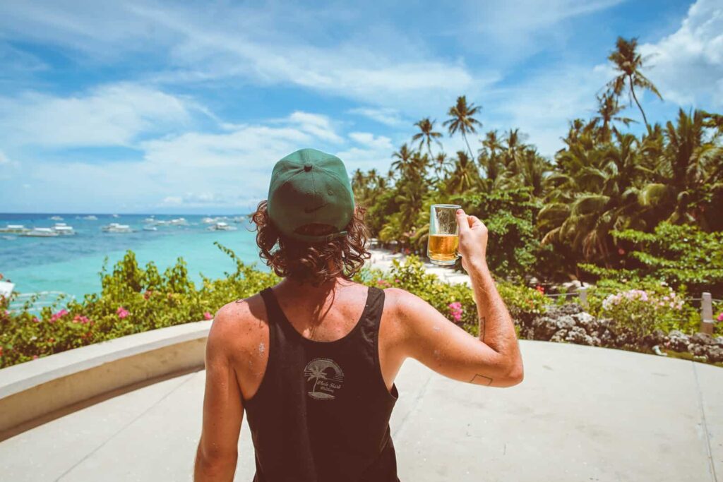 Back view of a person in a green cap and black tank top, raising a beer mug toward a tropical beach with palm trees and turquoise water.