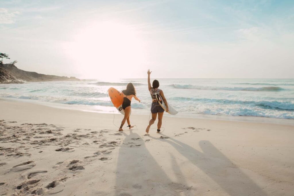 Two women surfers walk along a sandy beach toward the ocean, each carrying a surfboard as the sun sets behind them.