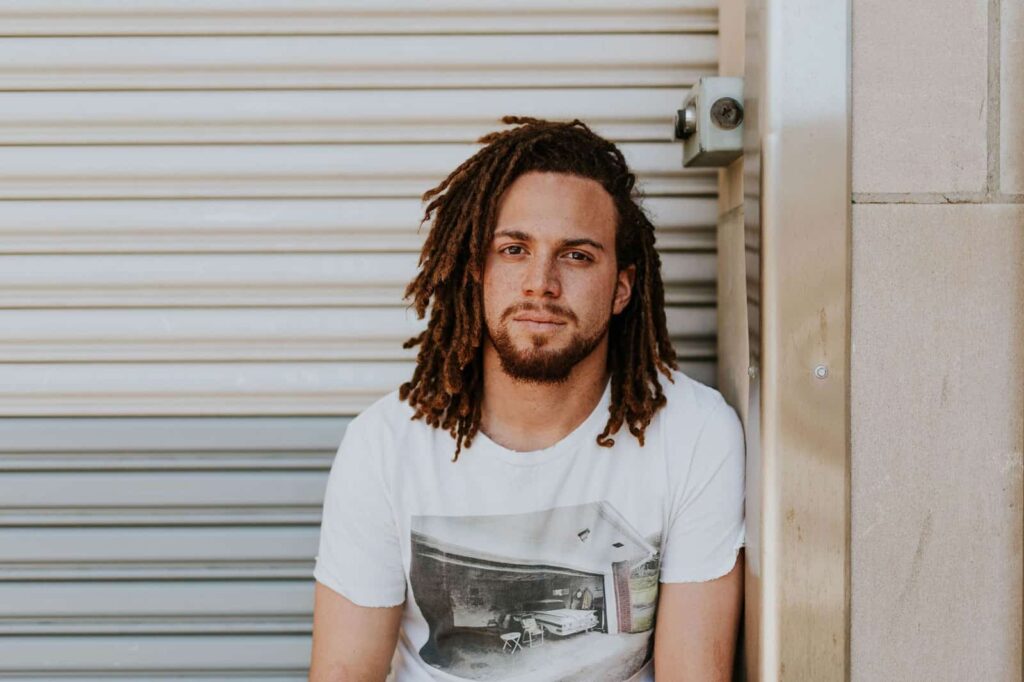 Portrait of a man with brown dreadlocks and a beard, wearing a white graphic t-shirt, leaning against a beige corrugated shutter outdoors.
