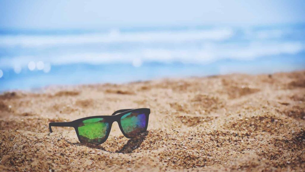 Black sunglasses with green mirrored lenses resting on the sandy beach, with the blue ocean in the background.