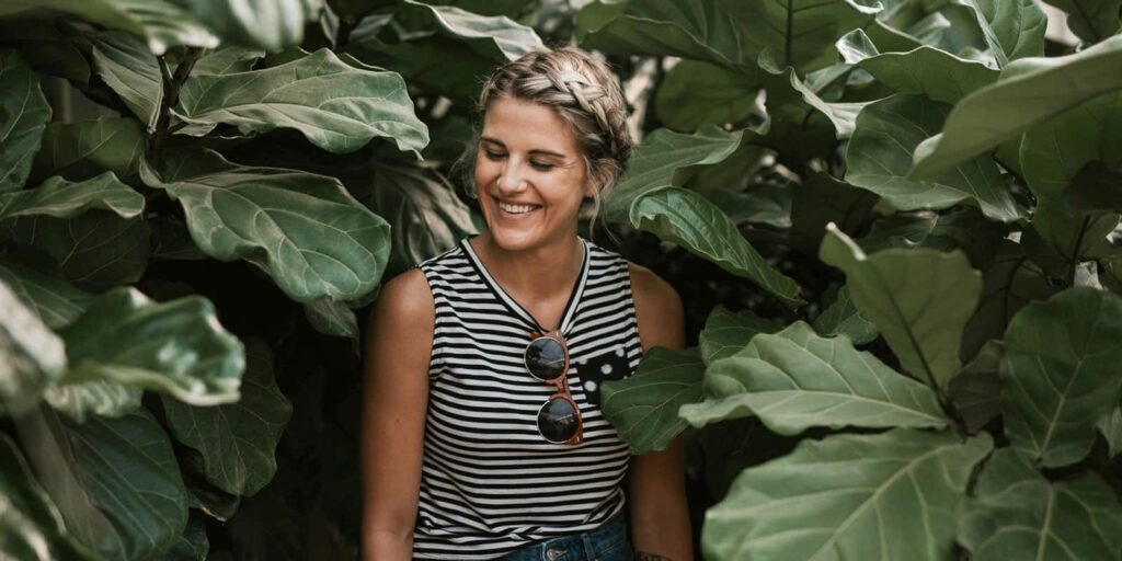 Smiling woman among large green leaves, wearing a black-and-white striped sleeveless top with sunglasses hanging from her neckline.