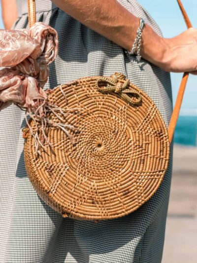 Person by the sea wearing a checkered dress, carrying a round woven straw bag with a pink scarf tied to the strap