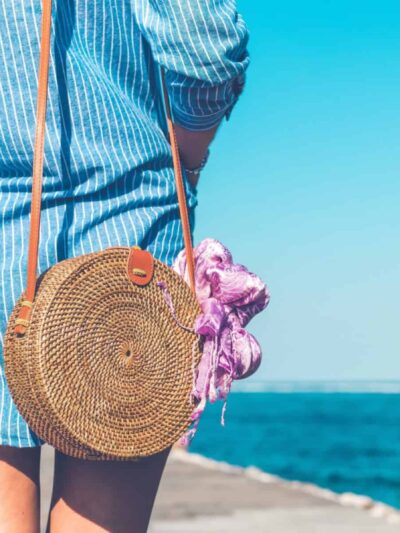 Person in a blue striped dress at the beach carrying a round woven straw bag with an orange strap and a pink scarf.