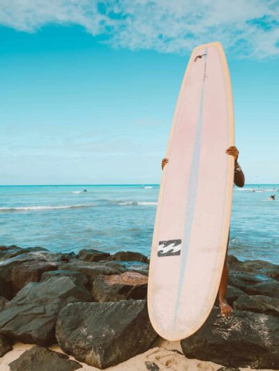 Person standing on rocky beach holding a tall pink surfboard upright near the water.