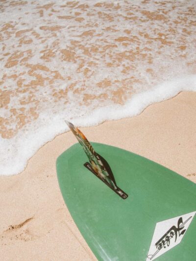 Green surfboard resting on sandy beach with a decorative fin and white foam from a receding wave nearby.