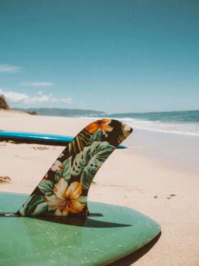 Green surfboard on sandy beach with a tropical floral-pattern fin sticking up from the board to the right of the frame.