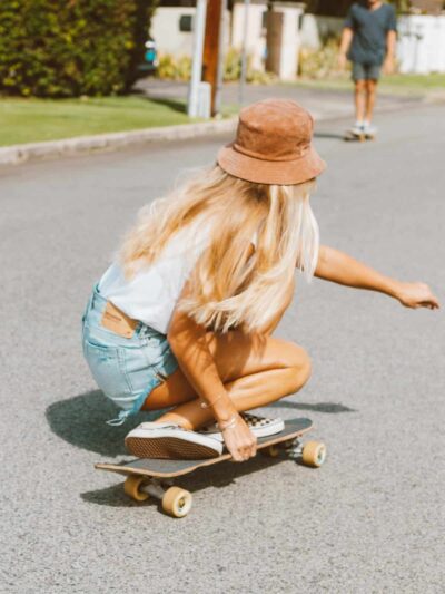 Woman with long blonde hair in a brown bucket hat crouches on a longboard, skating on a sunny residential street