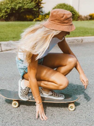 Person with long blonde hair and a brown bucket hat crouches on a skateboard on a sunny street.