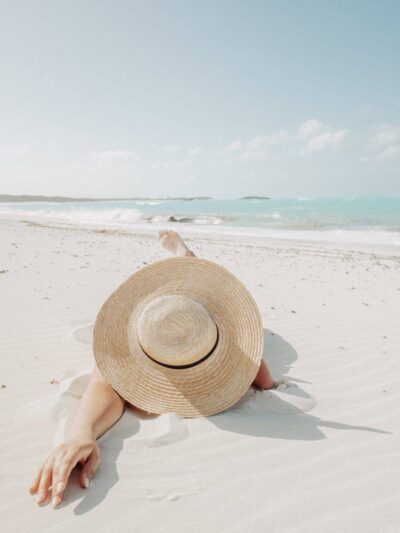 Person sunbathing on a white-sand beach with a large straw hat covering their face, turquoise ocean in the background.