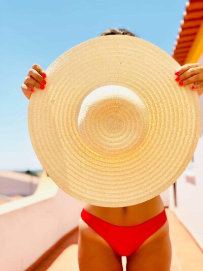 Person in a red bikini holds a large straw hat in front of their face on a sunny beach.