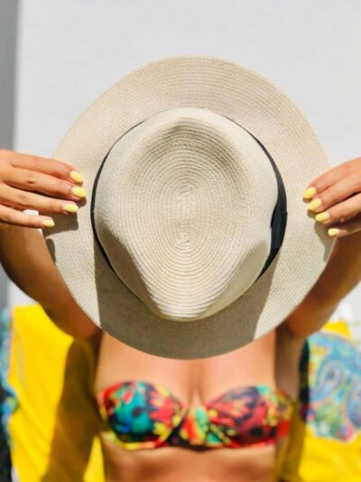 Person in a colorful bikini holds a large straw hat up to cover their face, showing yellow nail polish on both hands.
