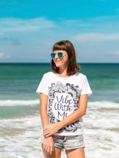 Woman standing on a sunny beach wearing a white graphic t‑shirt that reads 'Vibe With Me,' blue sunglasses, and shorts, with the sea in the background.
