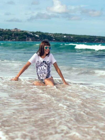 Woman kneeling in shallow surf at the beach, wearing a white shirt with 'Vibe With Me' and mirrored sunglasses, smiling toward the camera.