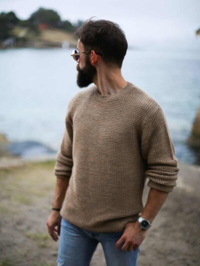 Man with a full beard and sunglasses stands by a body of water, wearing a tan chunky knit sweater and blue jeans.