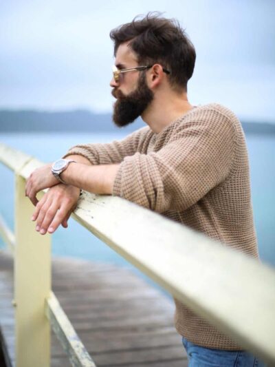 Man with a full beard and sunglasses leaning on a railing outdoors, wearing a beige knit sweater and jeans, gazing toward the water.