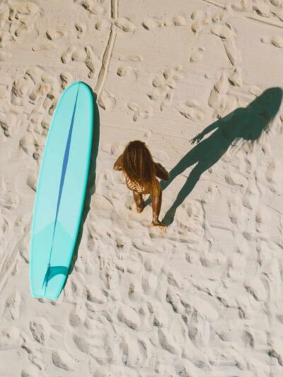 Overhead view of a person walking on pale sand beside a turquoise surfboard, with a long shadow extending to the right.