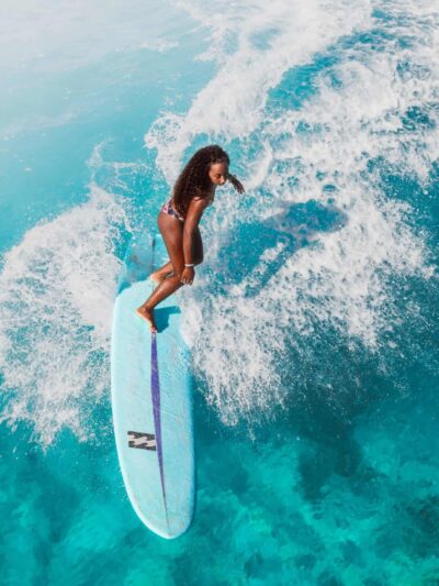 Female surfer balancing on a light blue surfboard as a turquoise wave crashes around her, spray filling the air