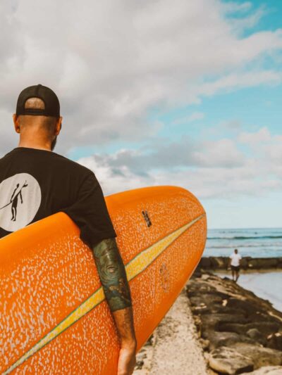 Surfer walking toward the sea with an orange surfboard under his arm, seen from behind on a rocky beach.