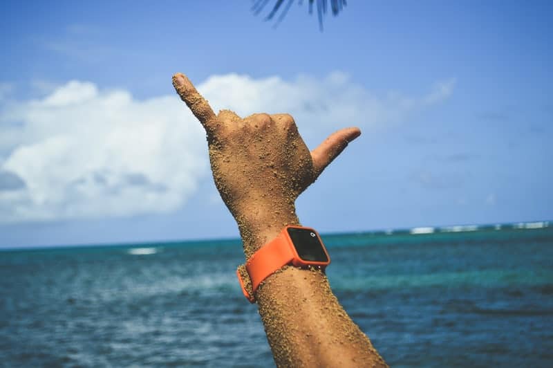 Arm raised at the beach, covered in sand, making a shaka (hang loose) gesture with an orange smartwatch on the wrist, blue ocean in the background.
