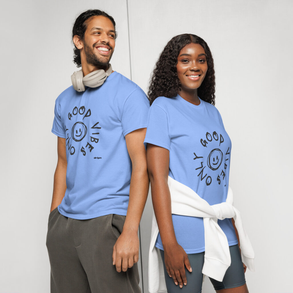 Two people in light blue t-shirts stand back-to-back, smiling at the camera; the man wears headphones around his neck against a neutral backdrop.